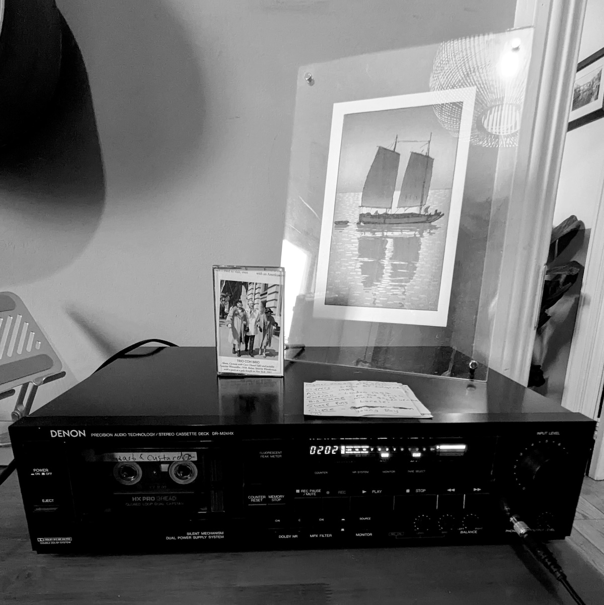 A Denon DR-M24HX cassette deck on a desk. On the deck is a cassette box with a cover made from a magazine cutout and a hand written track list as well as a woodblock print of Hiroshi Yoshida's Traditional Sailing Boat carved by David Bull.