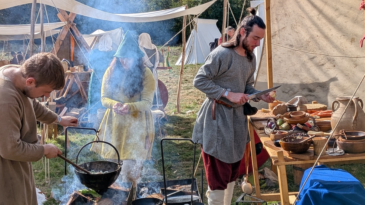 Three members of the Viking Re-enactment Society prepare and cook food over an open fire.